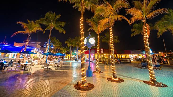 A clock pole in the middle of palm trees with lights, against outdoor dining at night
