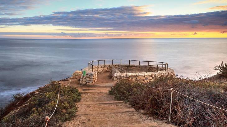 A walkway to a stone overlook against a sea-line under a partly cloudy sunset sky
