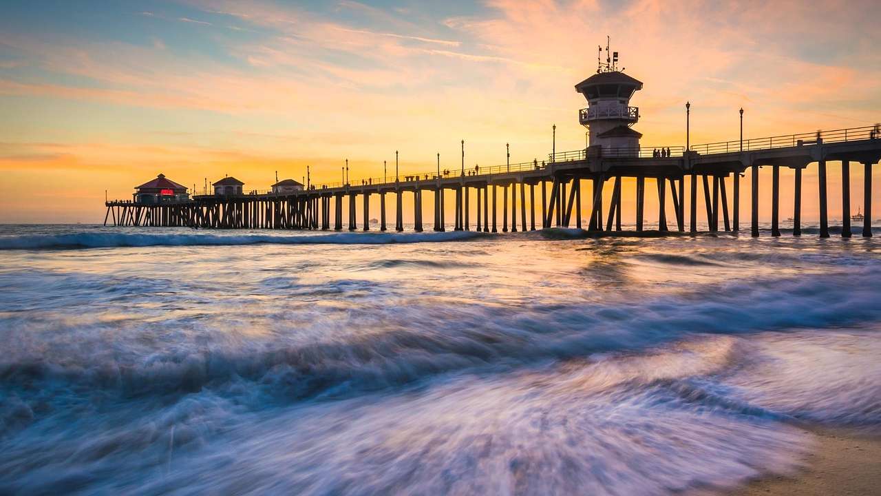 Looking towards a pier over water with waves, against a yellow-blue sunset sky