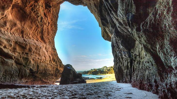 An open cave from which a sandy beach with seashore is visible under a blue sky
