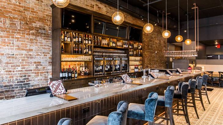 A wine bar with bar stools along the bar and bottles and a brick wall behind it