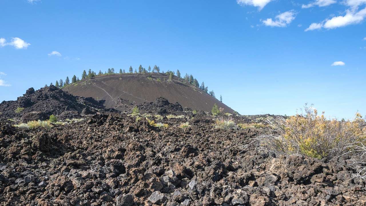 Looking at black volcanic rocks with some vegetation and a lava butte at the far back