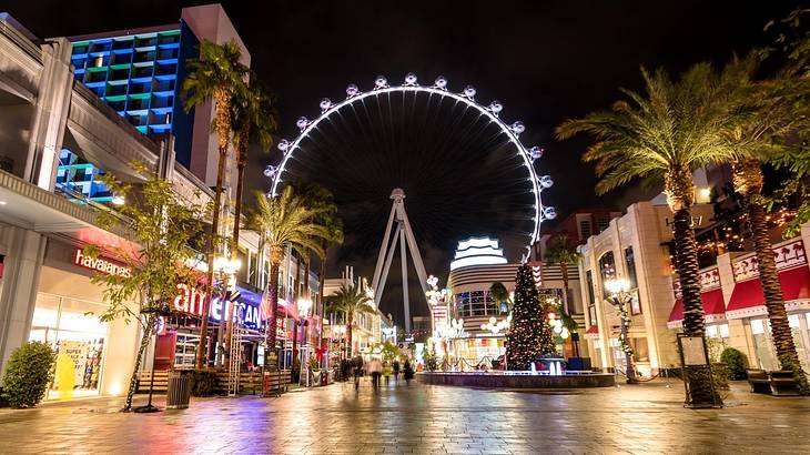 View of a Ferris wheel at night with buildings, a street, and palm trees in front