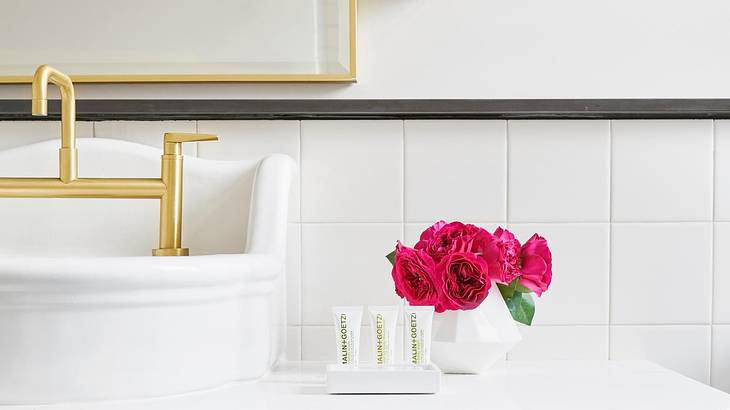 A bathroom with white tiles, a white sink and gold tap, and pink flowers