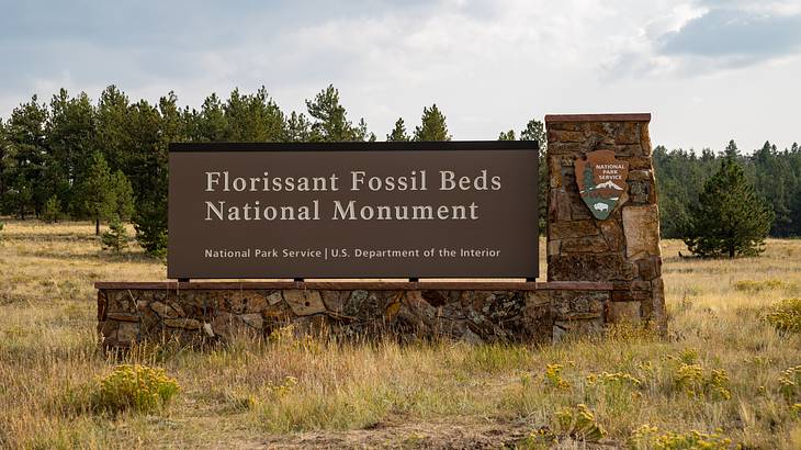 "Florissant Fossil Beds National Monument" signboard against trees & a cloudy sky