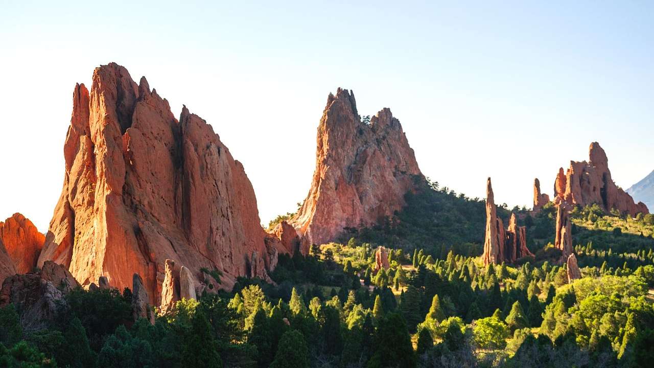 Several tall & massive red rock stacks surrounded by green trees under a clear sky