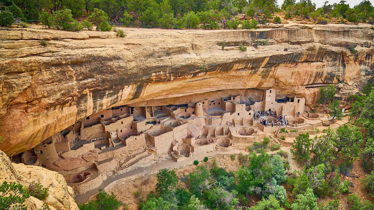 An aerial of a cliff with ancient dwellings and tourists gathered surrounded by trees