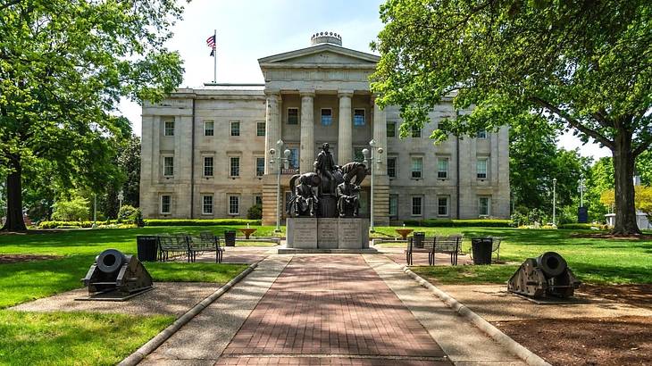 A stone building with columns and a path, statue, and canon in front of it A stone building with columns and a path, statue, and canon in front of it