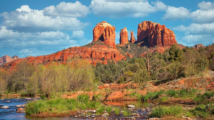 A tall red rock formation surrounded by vegetation & trees under a partly cloudy sky