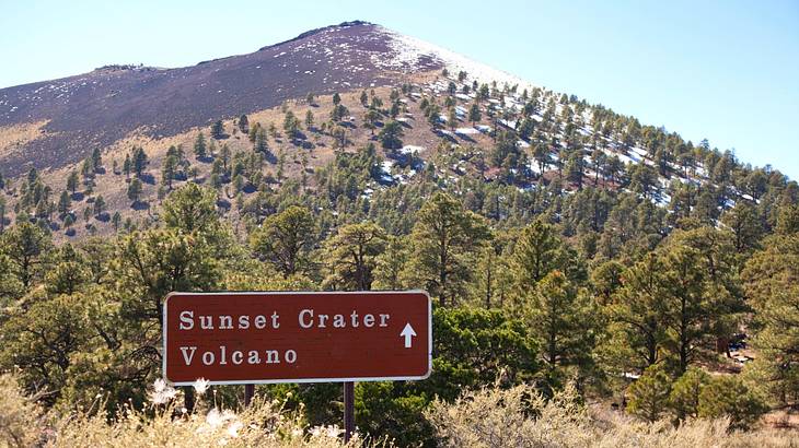A "Sunset Crater Volcano" sign against a mountain covered with trees and brown grass A "Sunset Crater Volcano" sign against a mountain covered with trees and brown grass