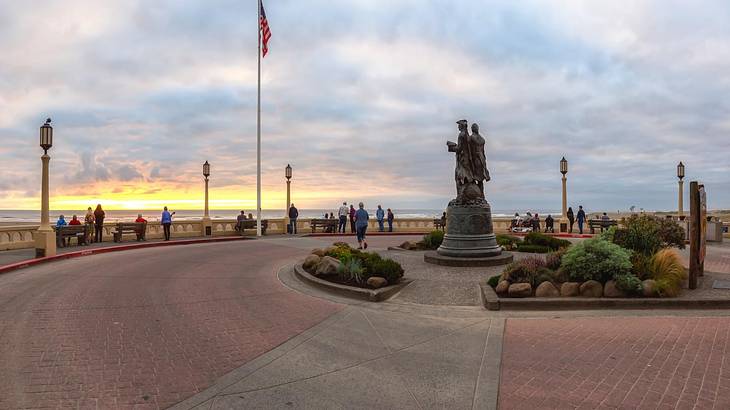 A beachside promenade with a flag, lamppost, and statue on it at sunset A beachside promenade with a flag, lamppost, and statue on it at sunset