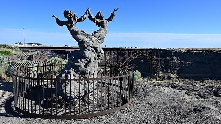 A grey statue of two women in front of a bridge and a canyon A grey statue of two women in front of a bridge and a canyon