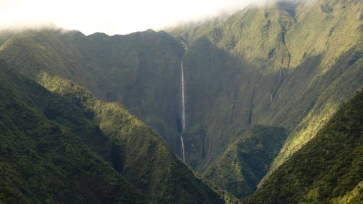 A tall and thin waterfall streaming through mountains covered in dense greenery