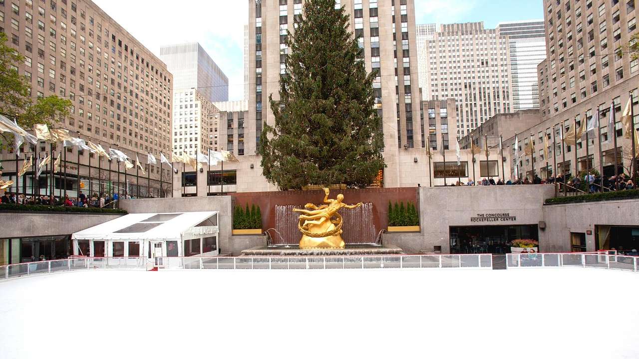 An ice rink with a gold statue and tree behind it and buildings surrounding it