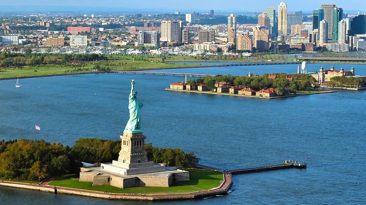 The Statue of Liberty surrounded by water with Ellis Island and a skyline behind it