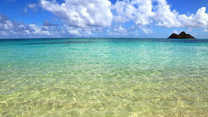 Clear ocean water with a hill in the distance under a blue sky with clouds