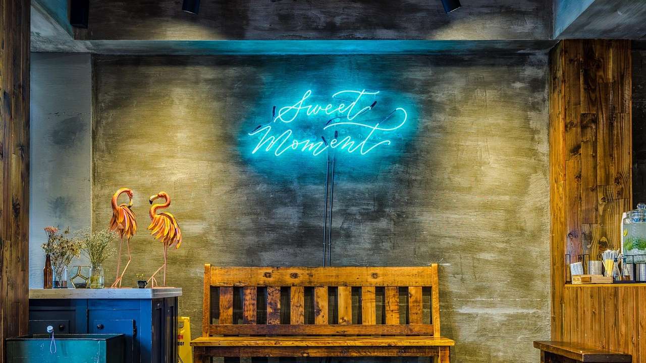 A cafe with a bench, flamingo sculptures, and a blue neon "Sweet Moment" sign