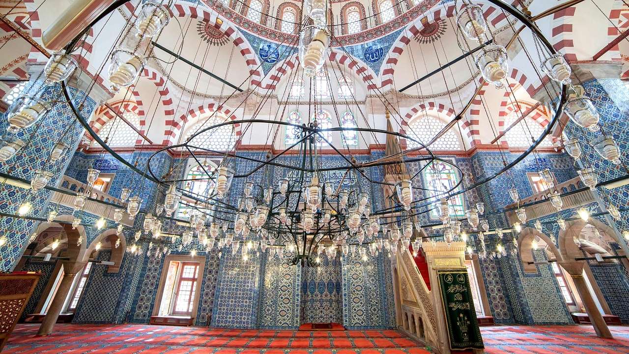 Inside a mosque with blue mosaic walls and a huge chandelier against a huge roof