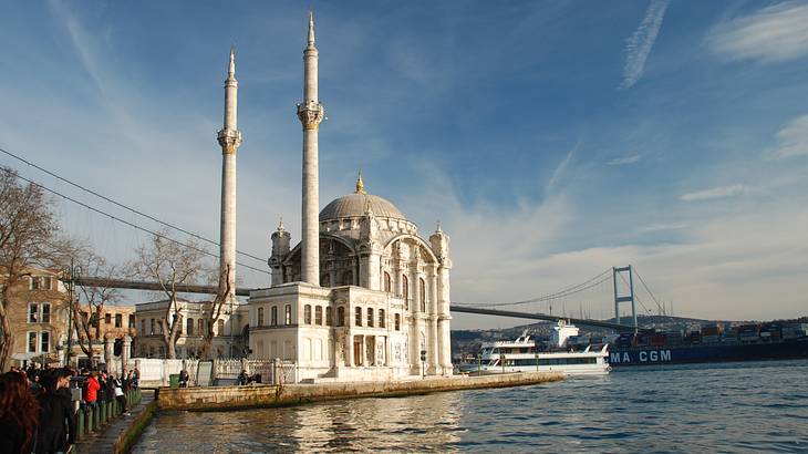 A white mosque on a pier next to a cruise ship and bridge over the water