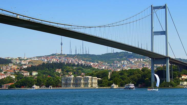 A suspension bridge over blue water against green trees and colorful buildings