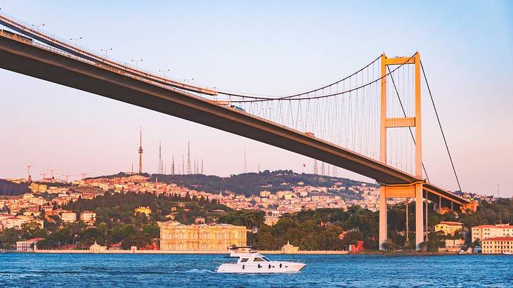 A suspension bridge over water with a cruise boat on it under an evening sky