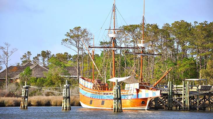 An old-fashioned ship sitting on the water with trees on the shore behind it