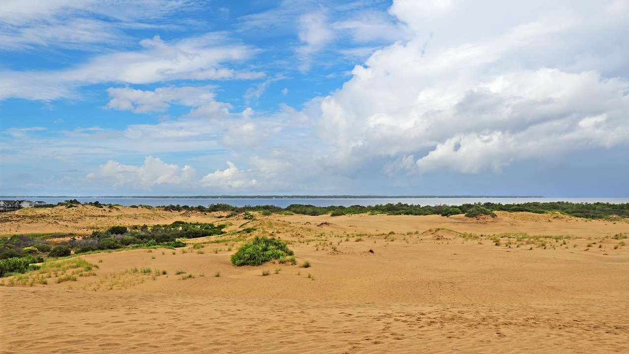 Sand with some greenery in it and water in the distance, under a blue sky with clouds