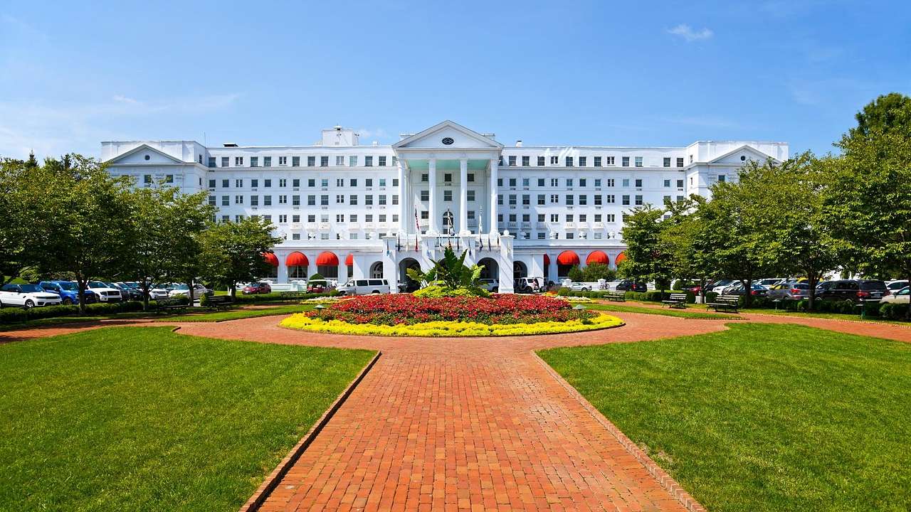 A white hotel building with columns and a path, grass, and colorful flowers in front