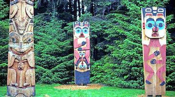 Three multi-colored totem poles with faces carved in them, in front of trees