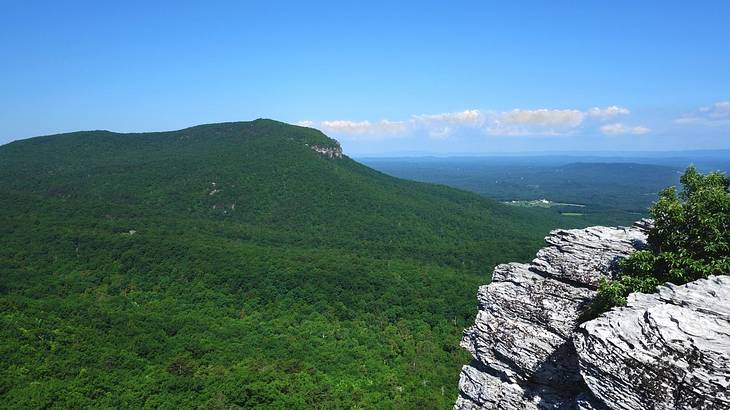 A greenery-covered hill with rocks to the side under a blue sky with some clouds