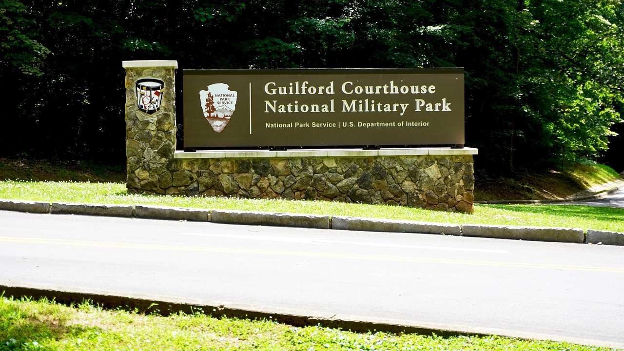 A Guilford Courthouse National Military Park sign next to grass, trees, and a road