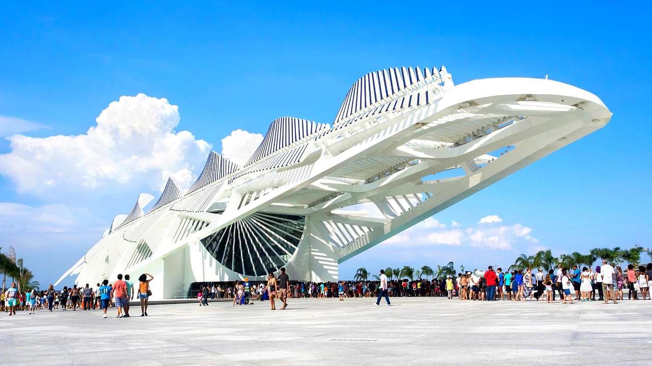 A white, tilted, futuristic structure surrounded by tourists and a blue sky