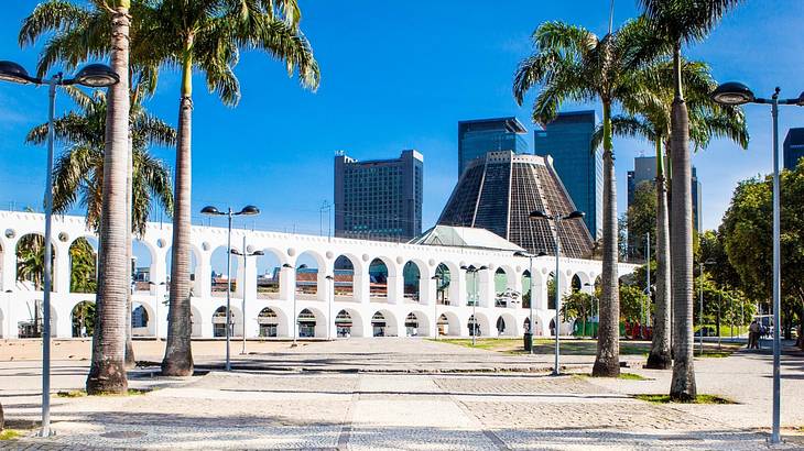 White arches with buildings behind them and palm trees and a street in front of them