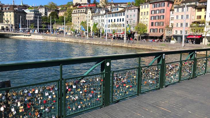 A green footbridge with padlocks and colorful buildings and a river behind it