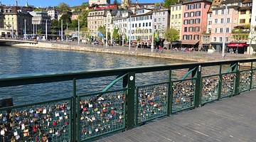 A green footbridge with padlocks and colorful buildings and a river behind it