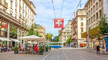 A tram track on a bustling road with shops and outdoor seating around it