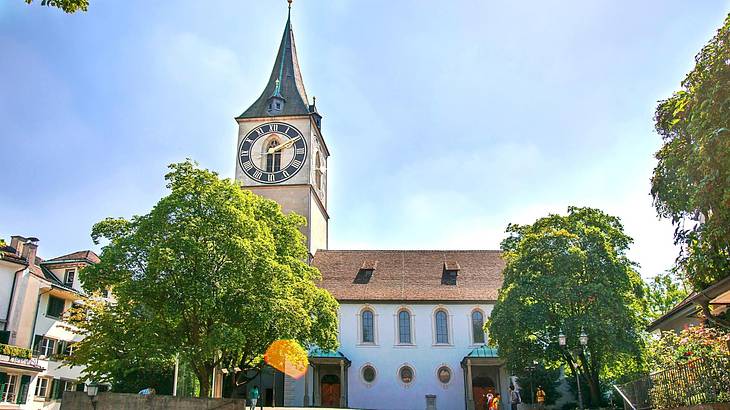 A white church with a tall clock tower surrounded by trees under a blue sky