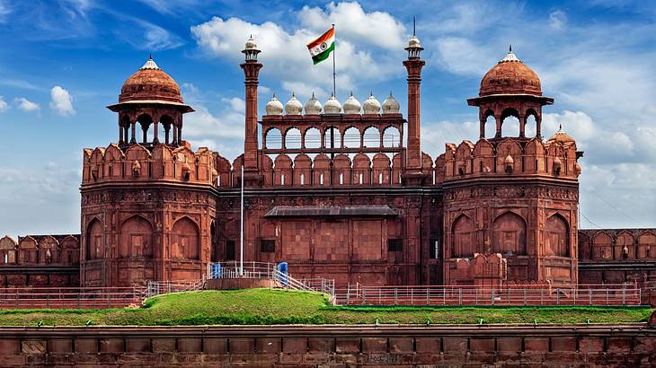 A red fort with two domed towers and an Indian flag against a partly cloudy sky