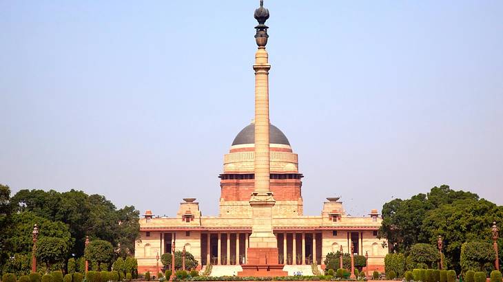 A tall sculpture in front of a neoclassical building with a dome surrounded by trees