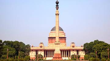 A tall sculpture in front of a neoclassical building with a dome surrounded by trees