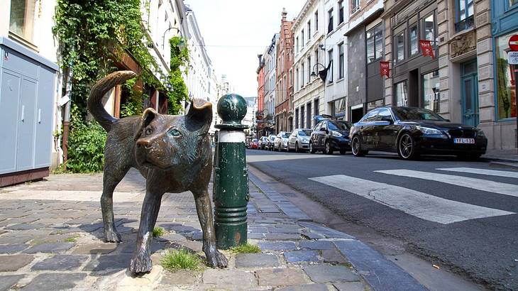 A bronze sculpture of a dog urinating on a sidewalk with cars and buildings opposite