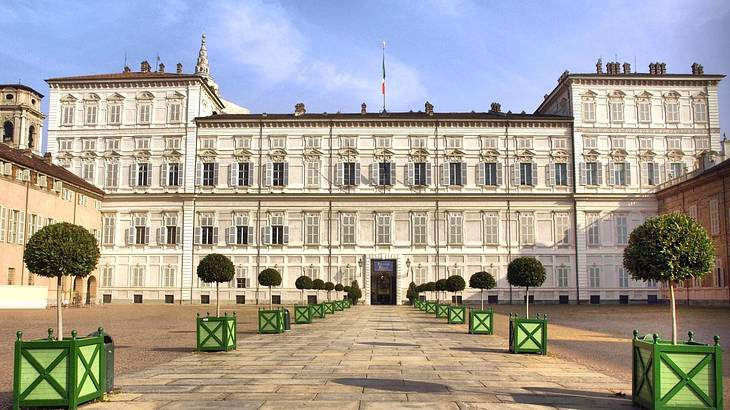 A courtyard with green bushes in green plant pots in front of a white palace building