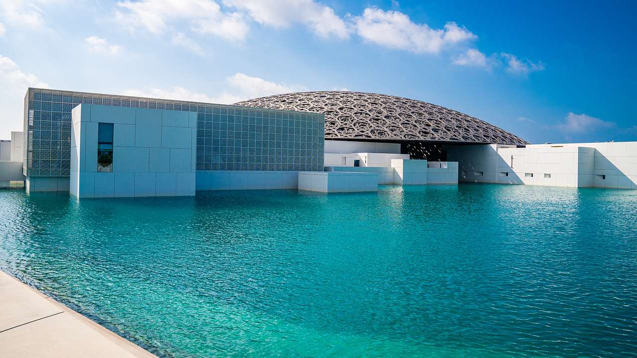 A contemporary white building with a round artistic gray roof next to a pool of water