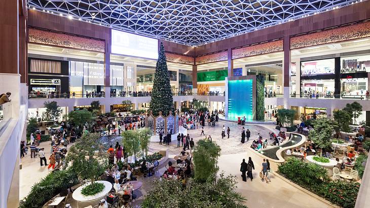 The interior of a mall with trees, people shopping, and a contemporary roof