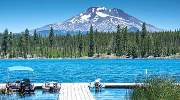 A dock on a blue lake against green pine trees and a snow-capped mountain
