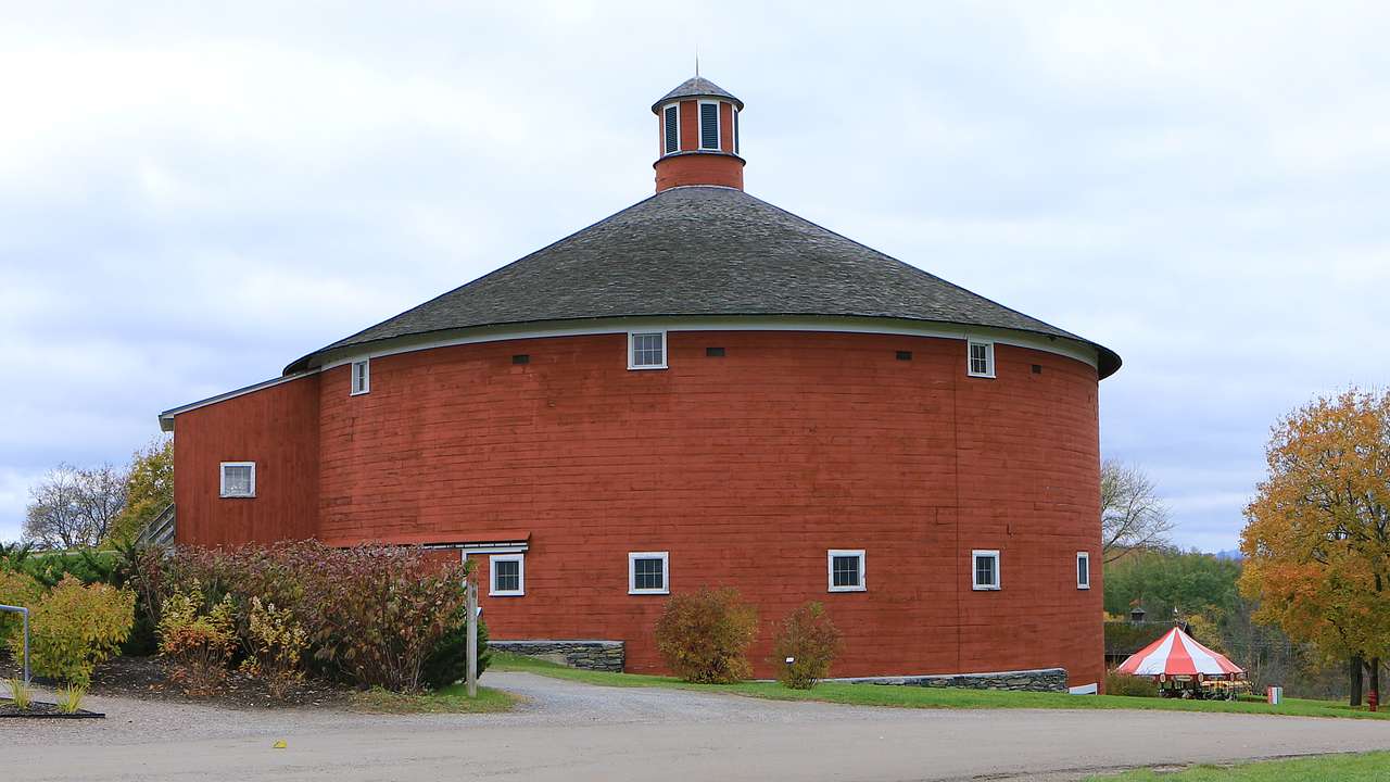 A red round barn with a paved road in front and bushes around