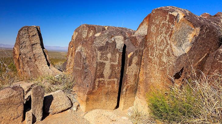 Historic petroglyphs carved onto huge rocks with some shrubs, under a clear blue sky