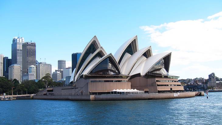 The outside of an opera house with white pointy tops along water and buildings