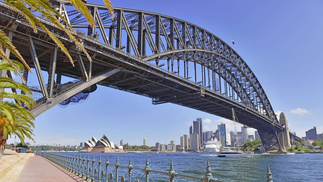 A steel arched bridge over water from below with a city skyline in the background