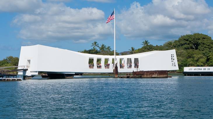 White memorial with a US flag on it along water, with trees and clouds in the back